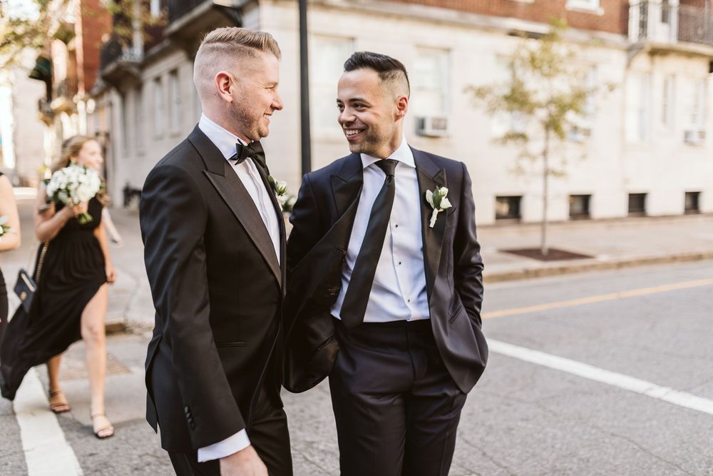 Two grooms in black tuxedos hold hands and smile while walking together on a city street on their wedding day.