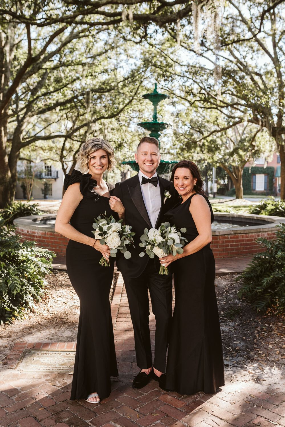Groom in tuxedo poses with two bridesmaids in black gowns holding white bouquets near a fountain.