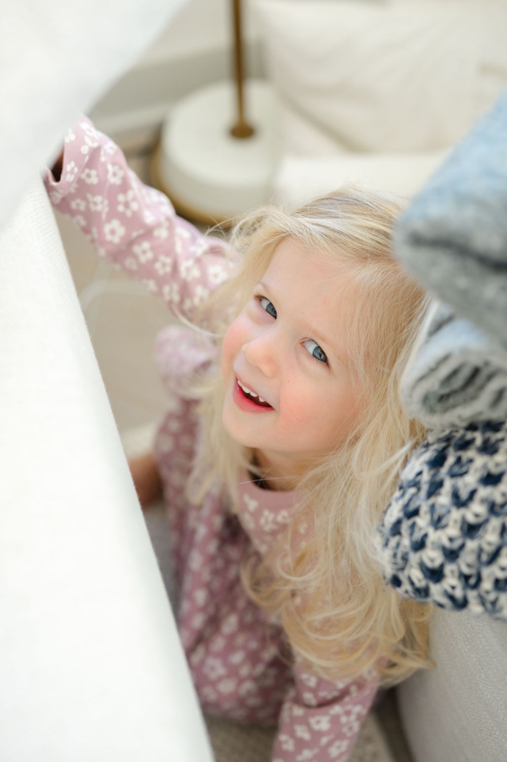 A smiling blonde toddler girl in pink floral pajamas reaches up while looking upward with bright blue eyes.