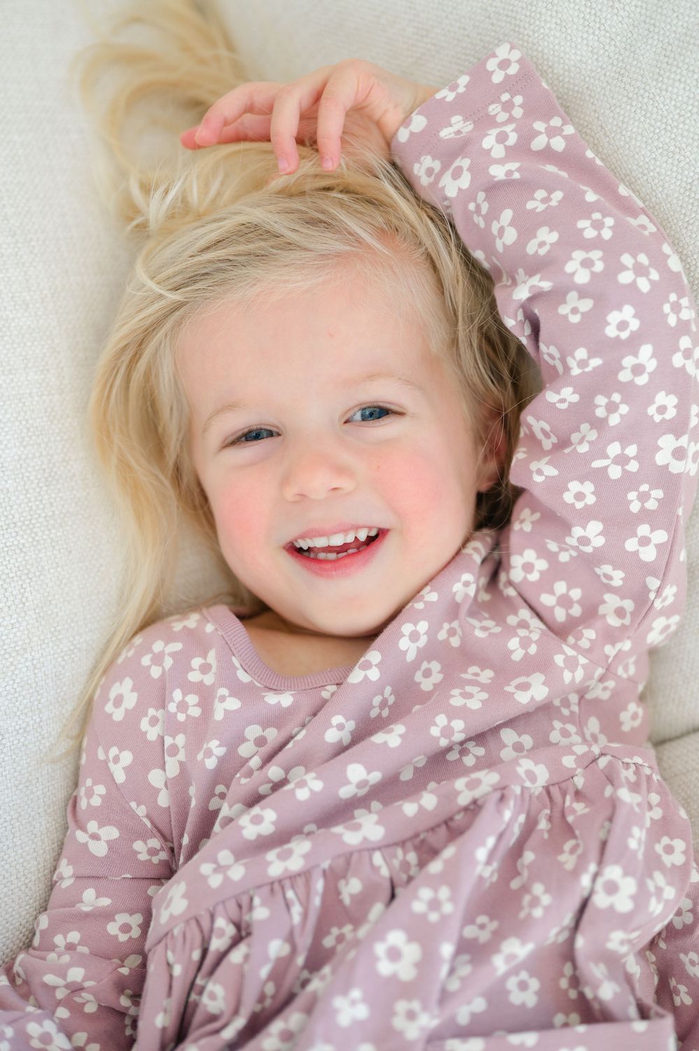 A cheerful blonde toddler girl in a pink floral dress laughing while lying on a white couch.