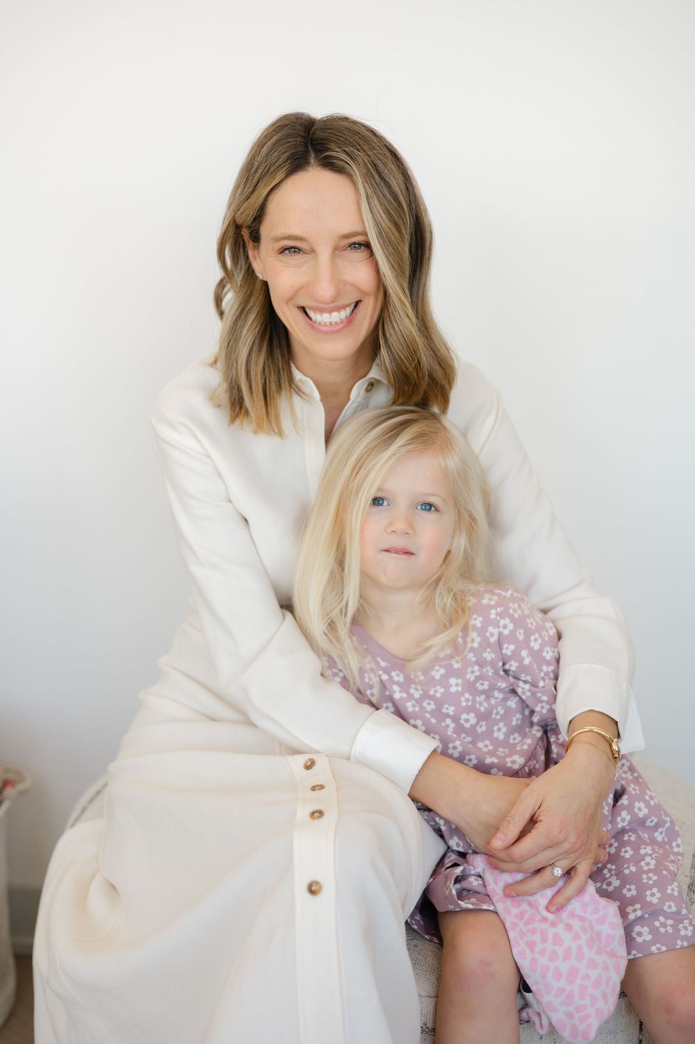 Smiling woman in white dress hugging young blonde girl in pink floral dress, sitting together against white background.