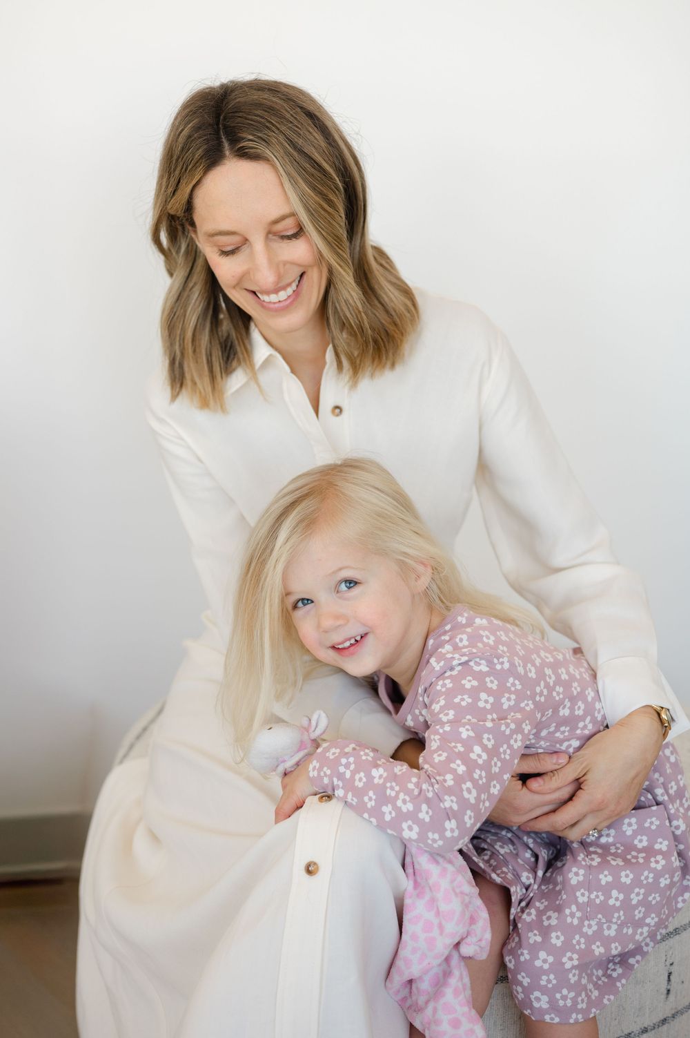 A smiling mother in a white dress lovingly embraces her young blonde daughter in a floral outfit.