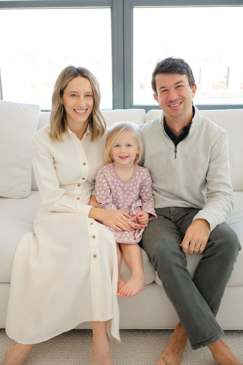A smiling family of three sits on a white sofa, with parents flanking a young blonde girl in a pink floral dress.
