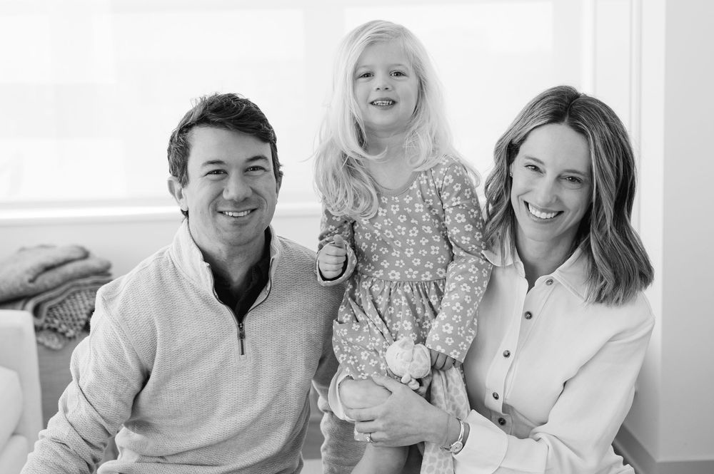 Black and white family portrait of smiling parents with young daughter in floral dress posing together indoors.