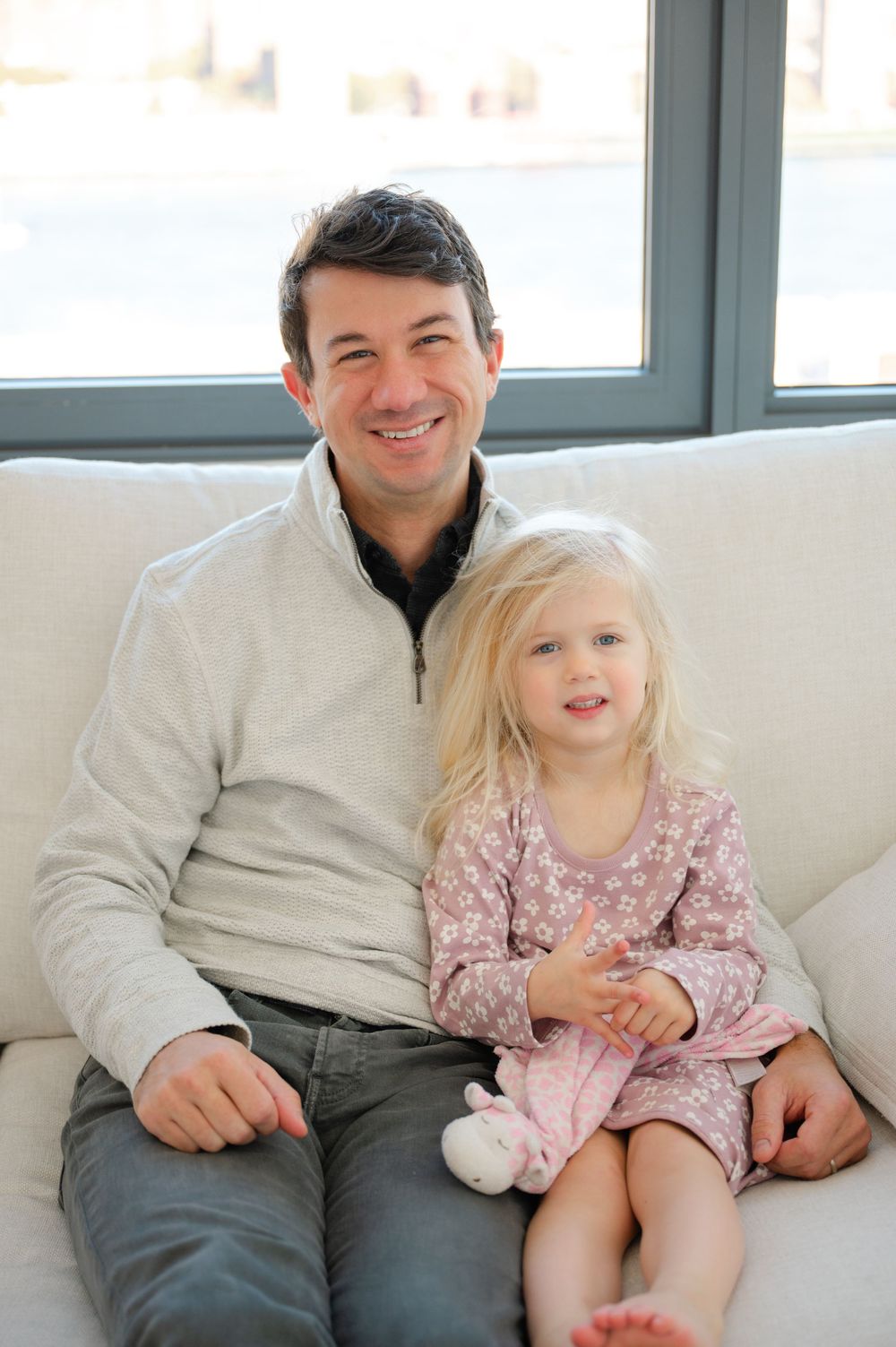 Smiling father in a gray pullover sitting on a white couch with his young blonde daughter in a pink floral dress.
