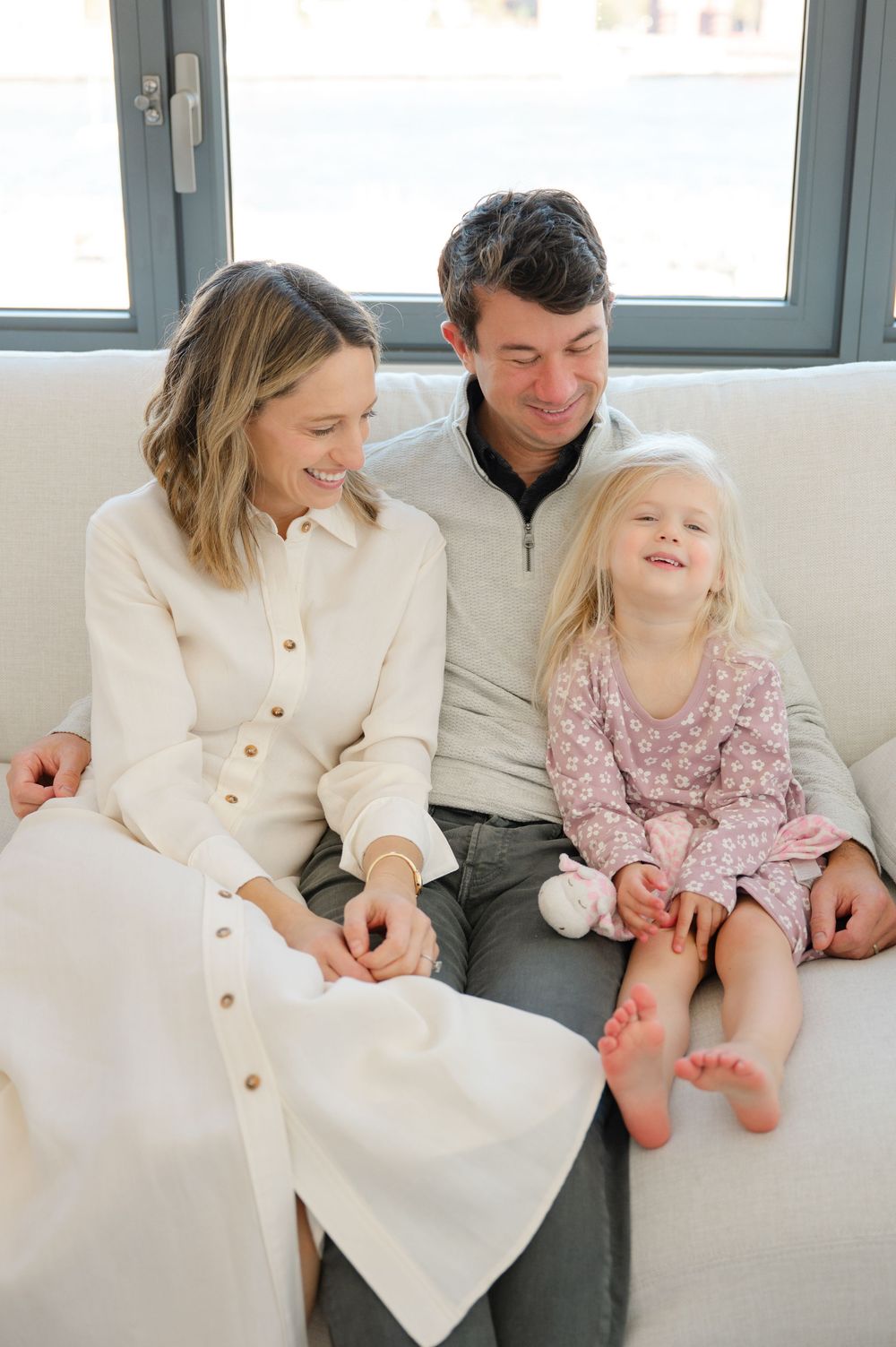 Happy family of three sitting on a white sofa, parents smiling at their young blonde daughter in a pink floral dress.