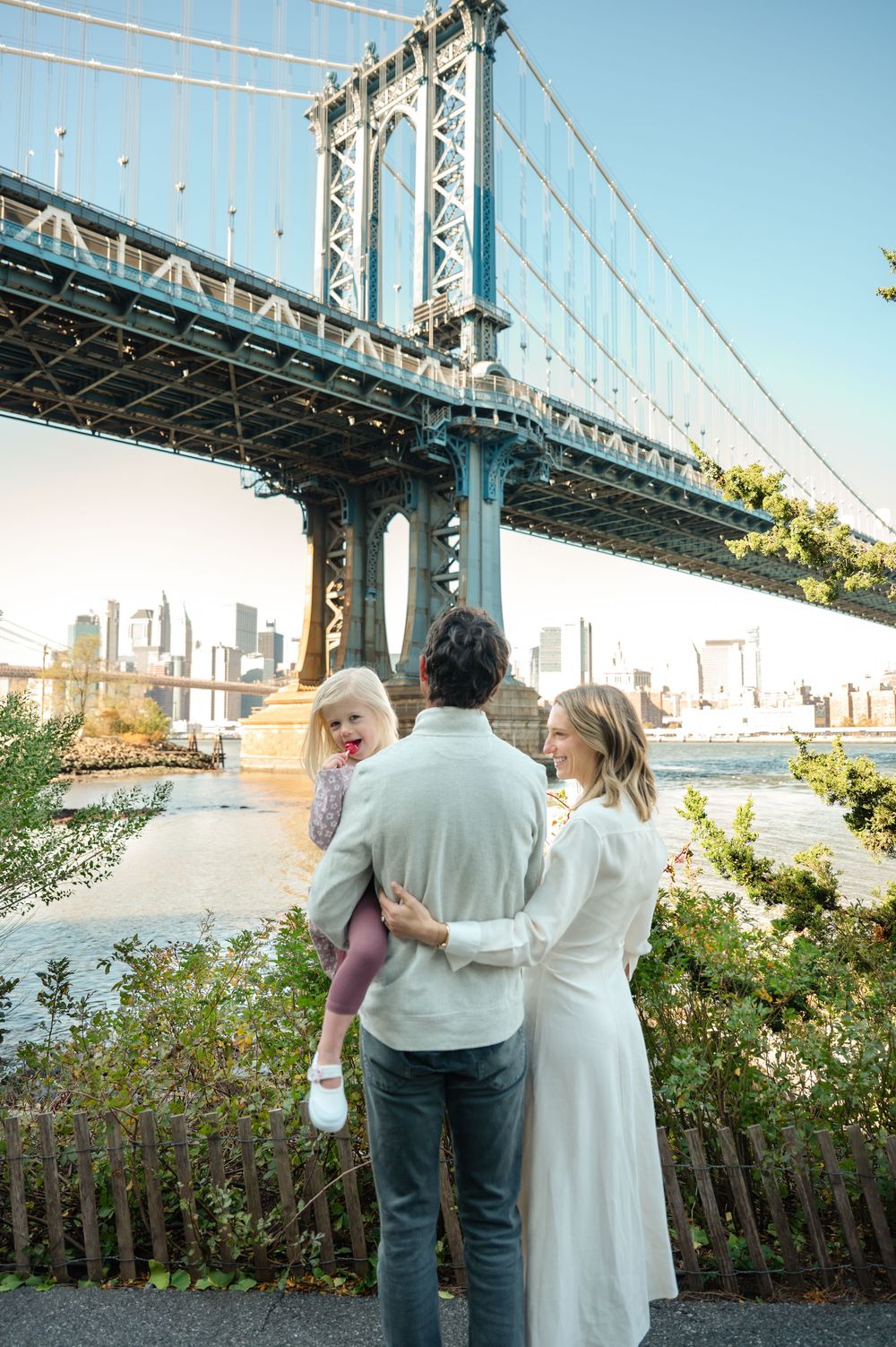 Family of three admiring the Manhattan Bridge from Brooklyn waterfront park with NYC skyline views.