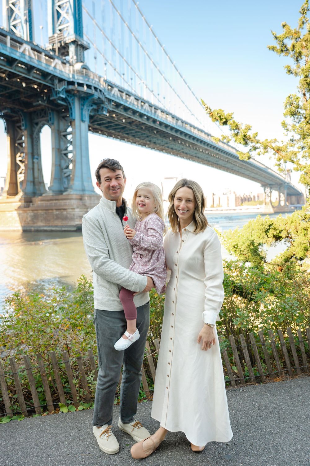 Family of three posing near Manhattan Bridge in NYC, man holding toddler girl, woman in white dress, sunny day.
