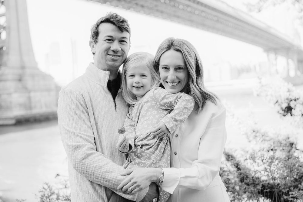 A happy family of three poses together outdoors near a bridge in a black and white portrait photo.