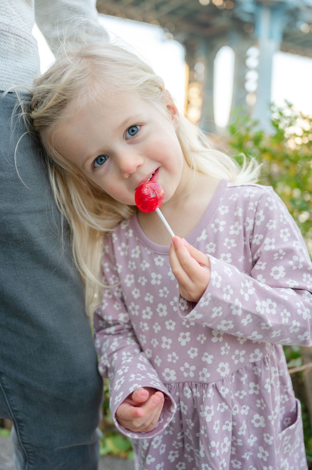 Blonde blue-eyed little girl in floral dress smiling while licking a red lollipop outdoors.