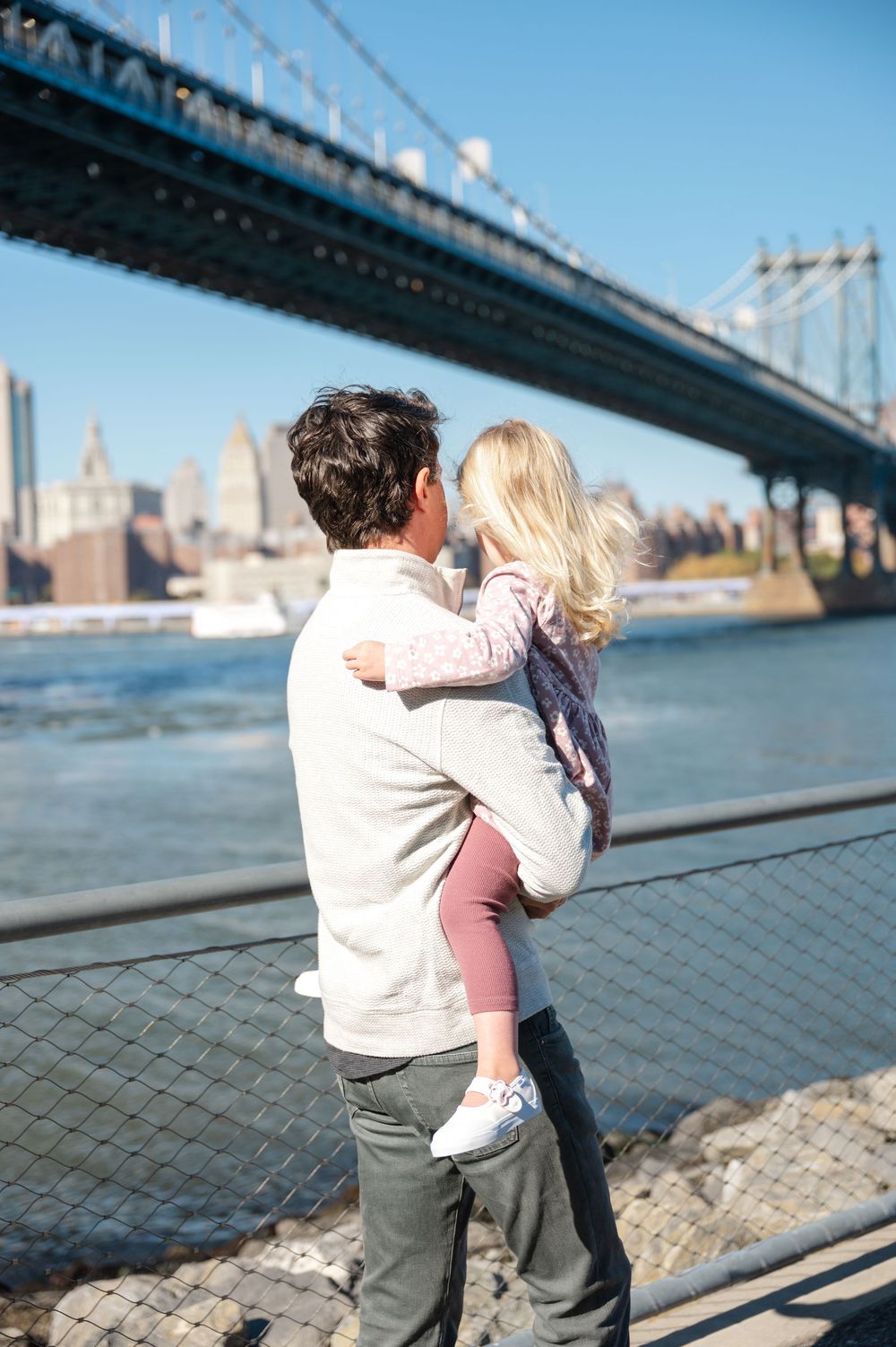 A parent holds a young child while standing by the waterfront with a large bridge and city skyline in the background.