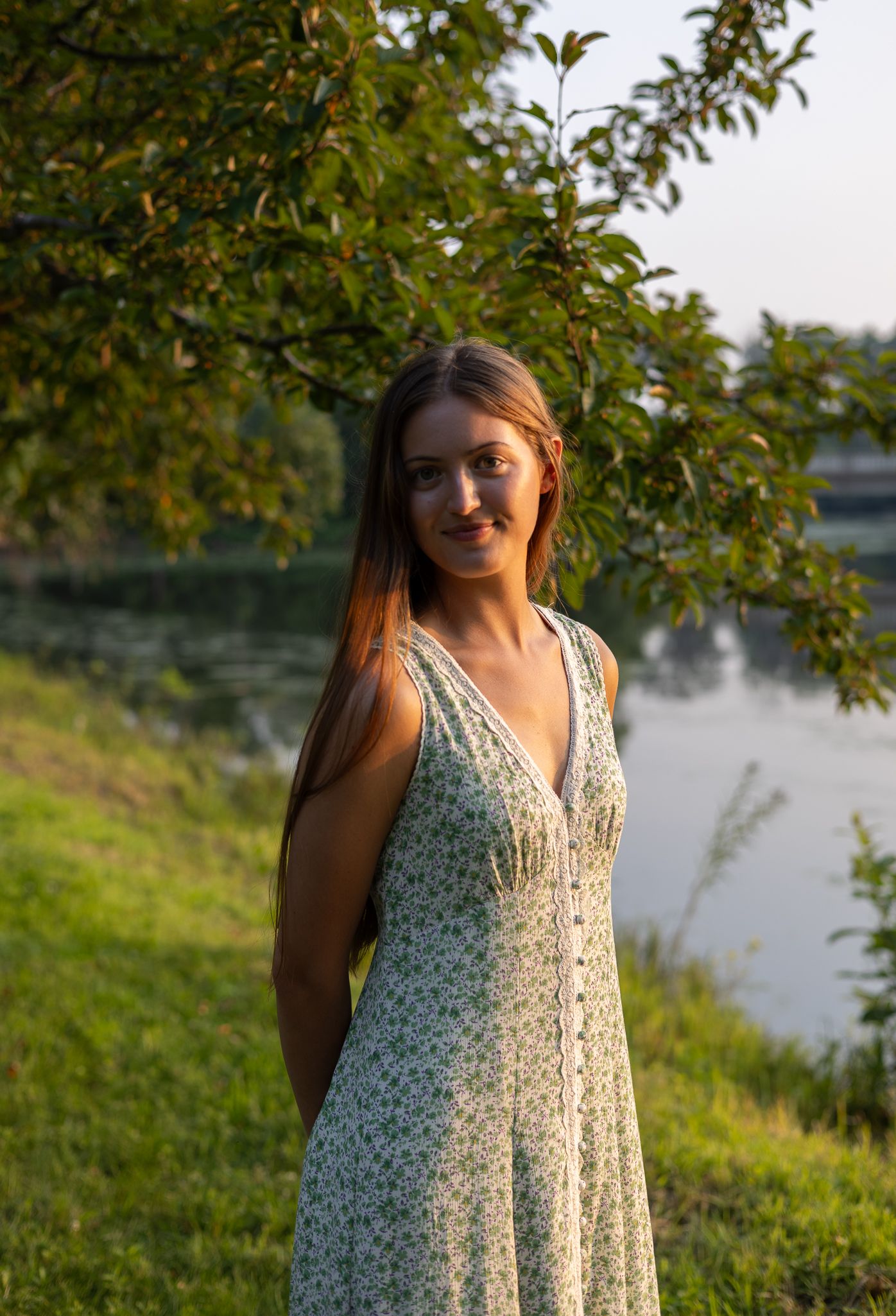 Young woman in floral sundress standing by a river under a leafy tree at sunset.