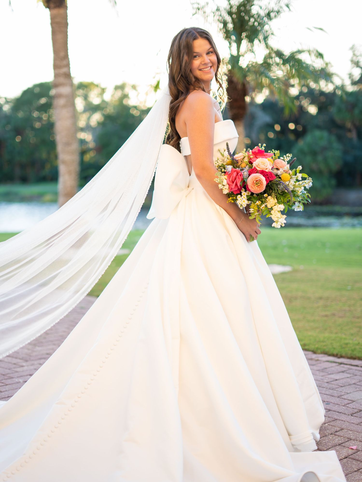 Bride in strapless ball gown with flowing veil holds colorful bouquet outdoors on brick path with lush greenery.