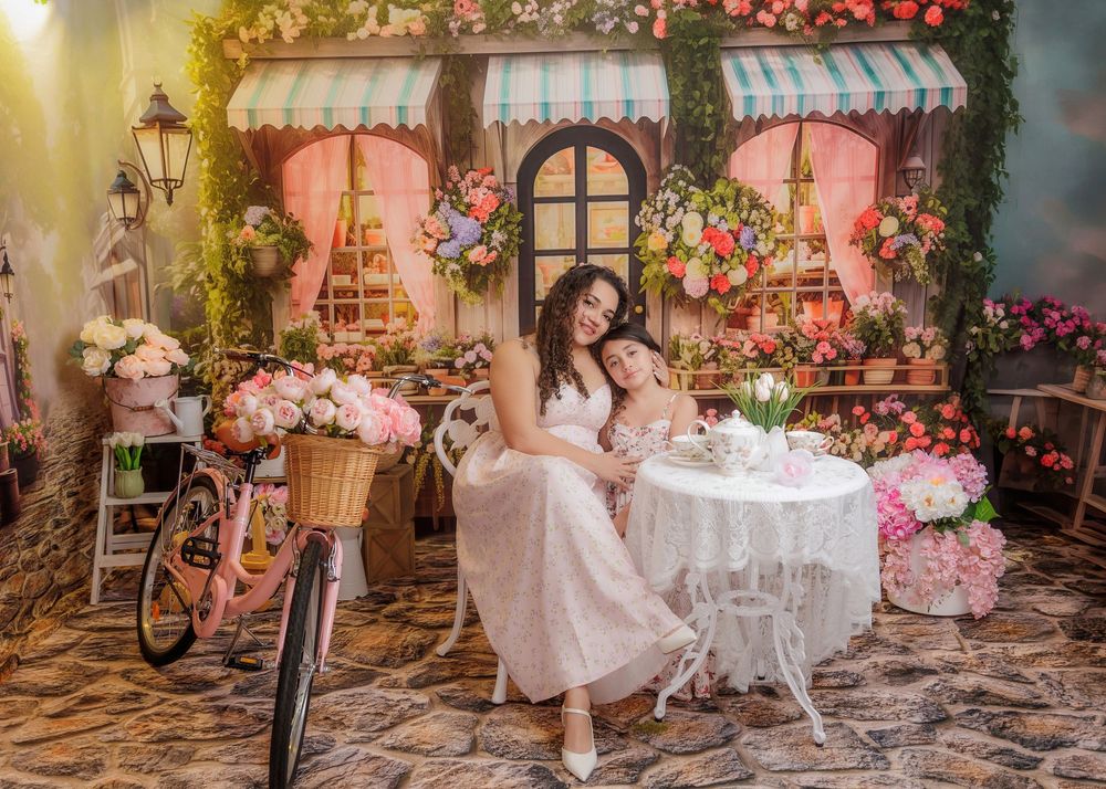 Mother and daughter in white dresses sit at a table beside a pink bicycle in a floral shop setting.