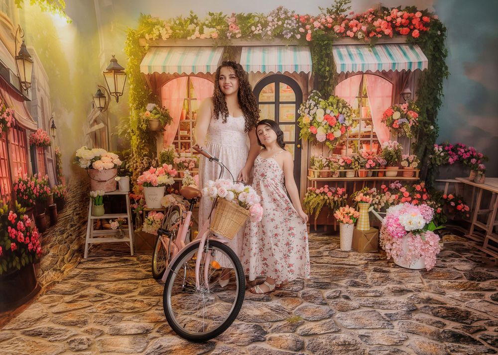 Mother and daughter in white and floral dresses pose beside a bicycle in a charming floral shop backdrop.