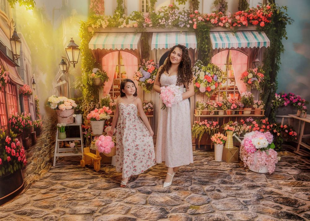 Mother and daughter in white dresses walk hand-in-hand through a whimsical floral shop backdrop.