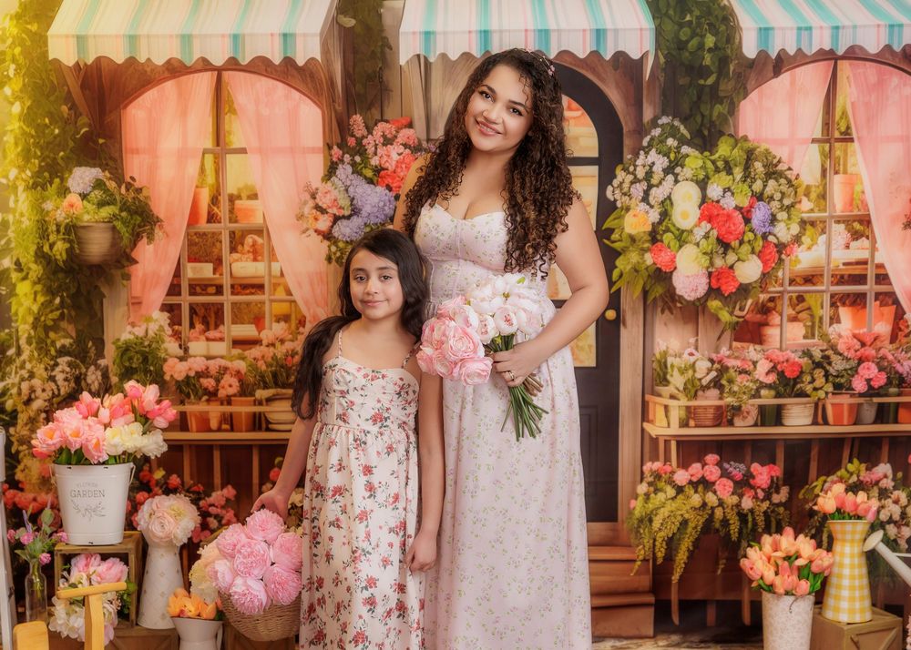 Mother and daughter in floral dresses pose together holding pink peonies in a vibrant flower shop set.