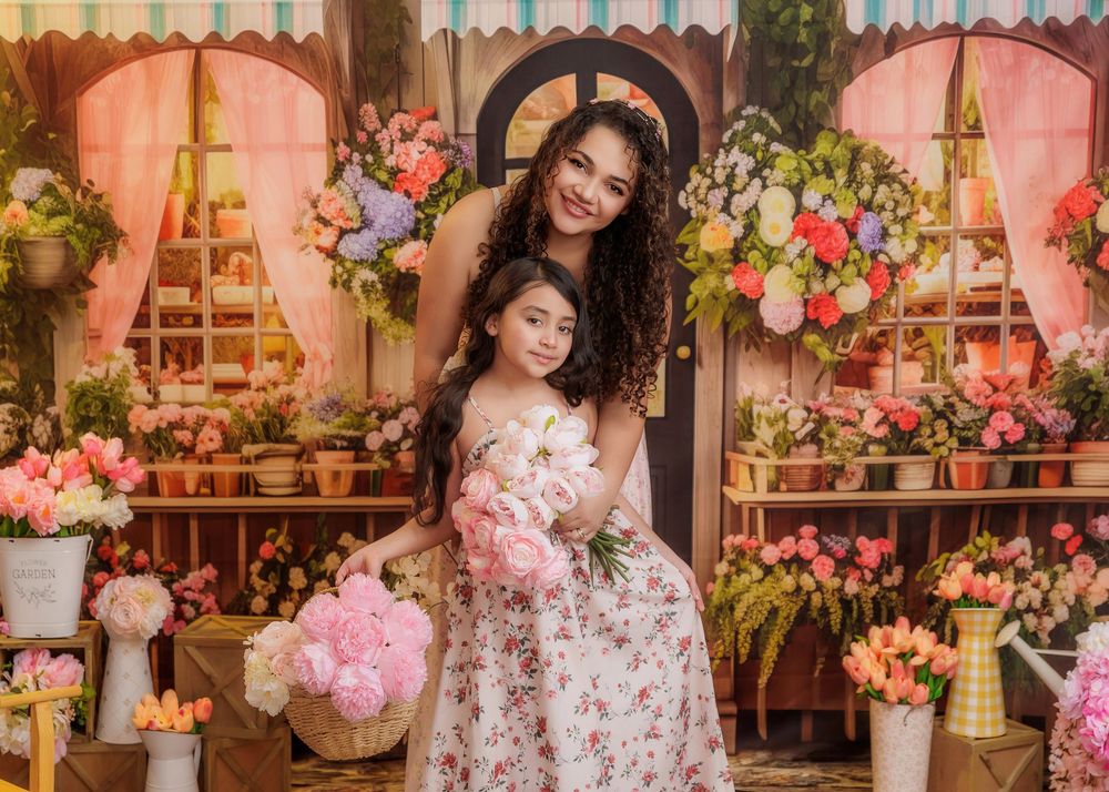 Mother hugs daughter in matching floral dresses while holding white lilies in a colorful flower shop set.
