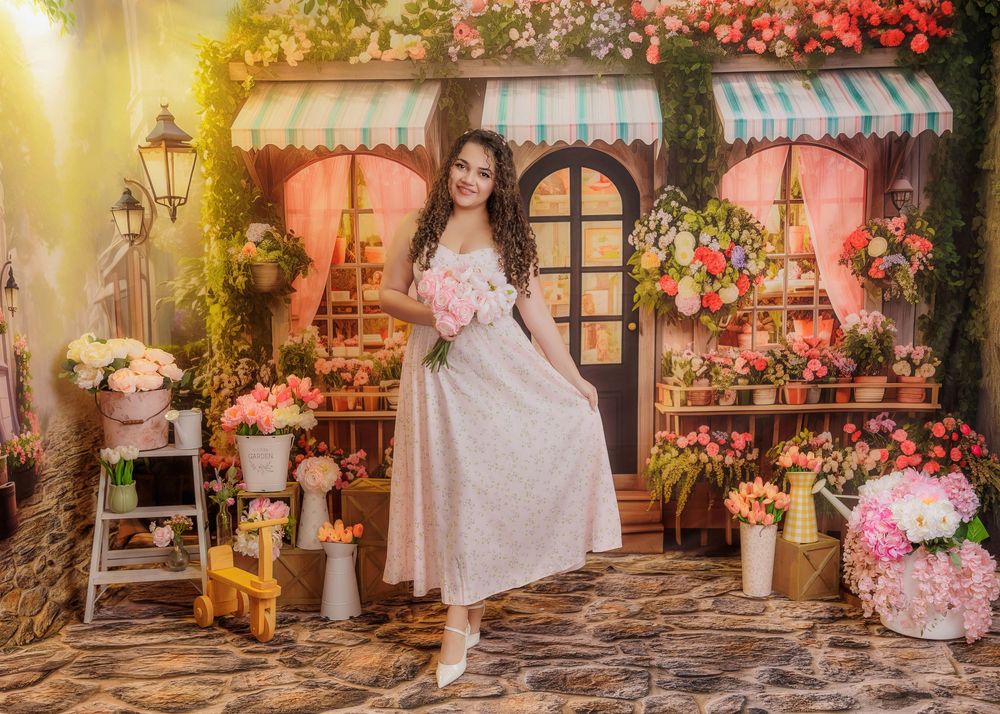 Woman in pink dress holding flowers poses in a charming floral shop backdrop with colorful blooms.