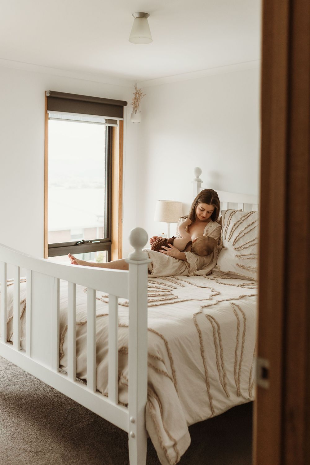 Mother breastfeeding newborn baby in bed with neutral beige bedding in a bright, cozy bedroom.