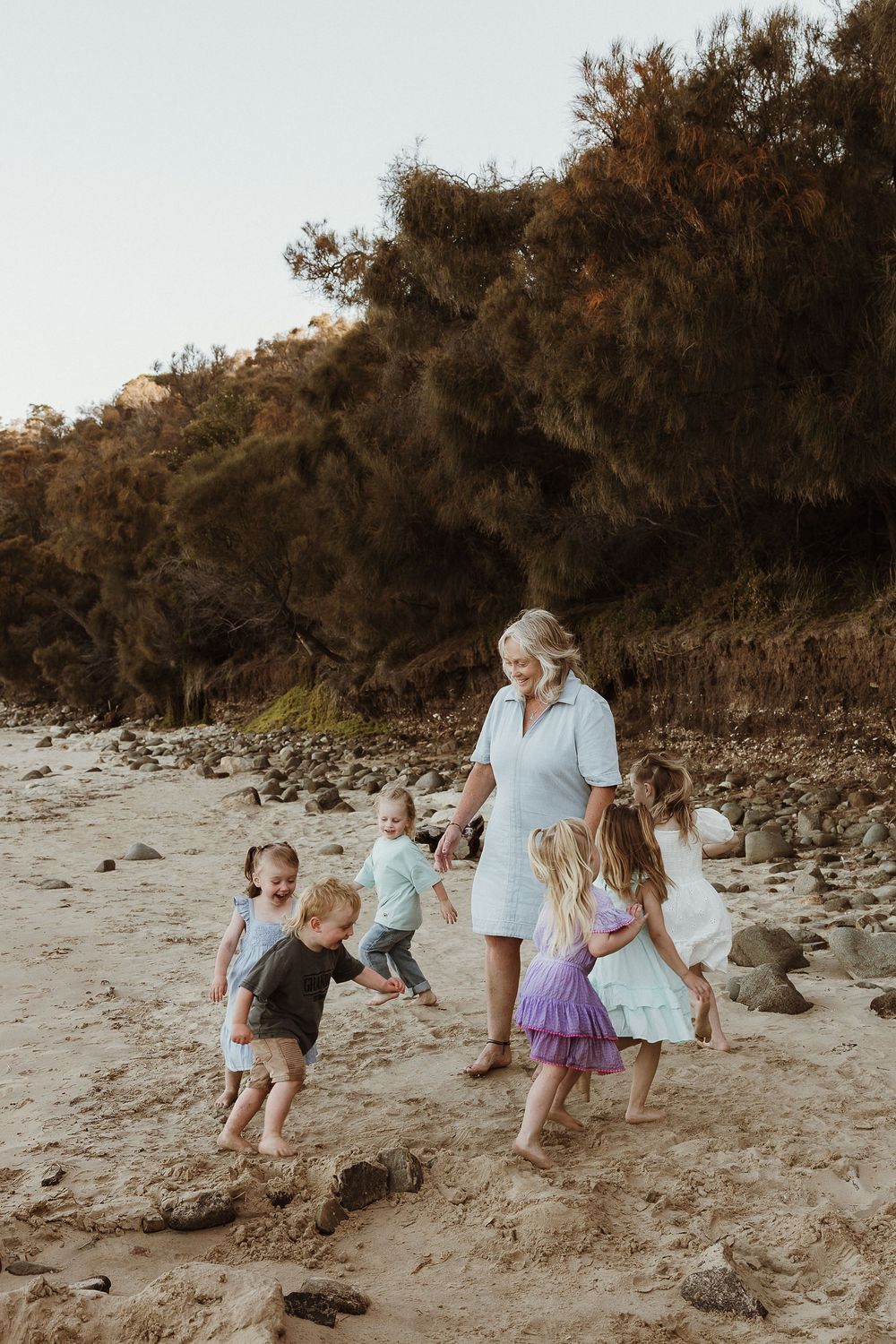 Grandmother playing with grandchildren on a sandy beach with rocky cliffs and trees in the background.