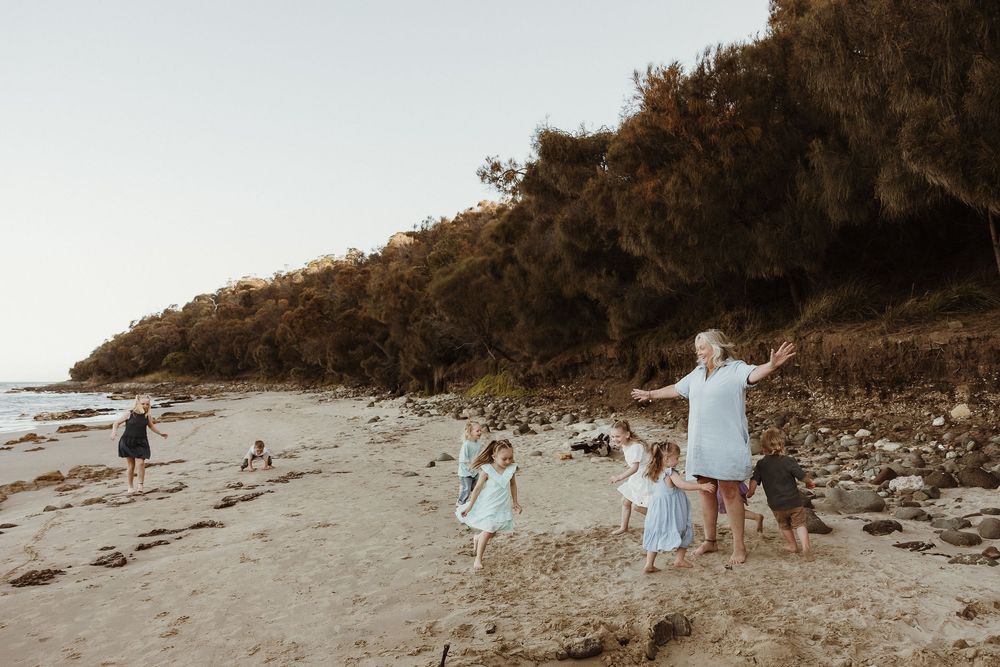 Family playing on a rocky beach with children and grandmother dancing near a tree-covered cliff at sunset.