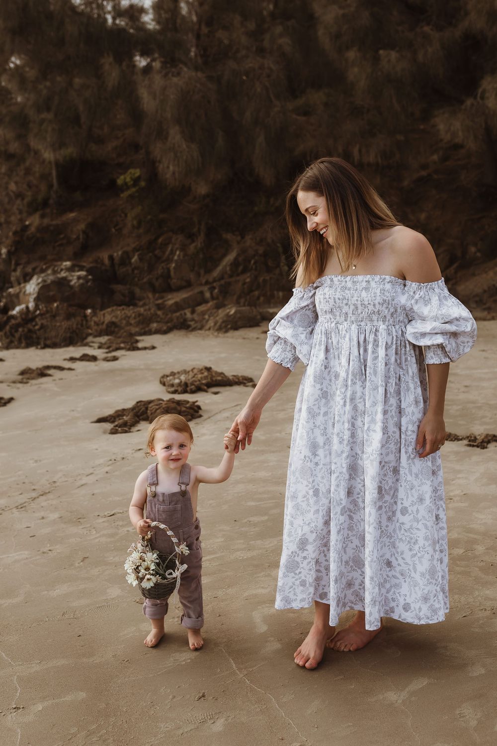 A mother in a floral off-shoulder dress holds hands with her toddler in overalls on a sandy beach.