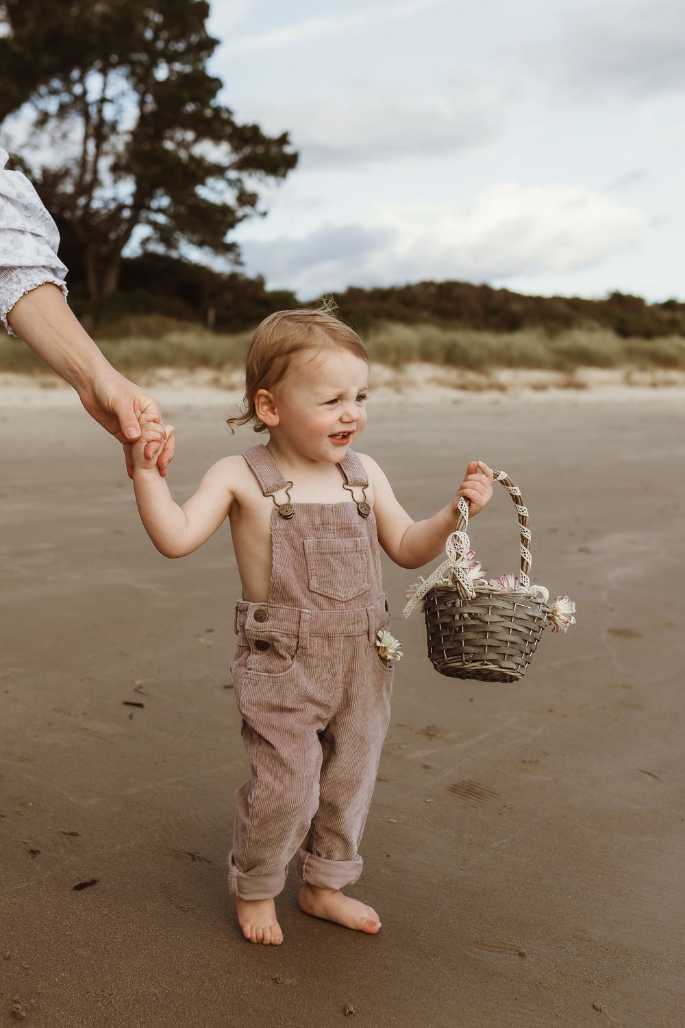 Toddler in beige overalls holding a wicker basket on a sandy beach, smiling while holding an adult's hand.