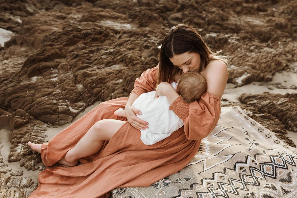 A mother in a rust dress tenderly holds her baby on a patterned blanket against rocky terrain.
