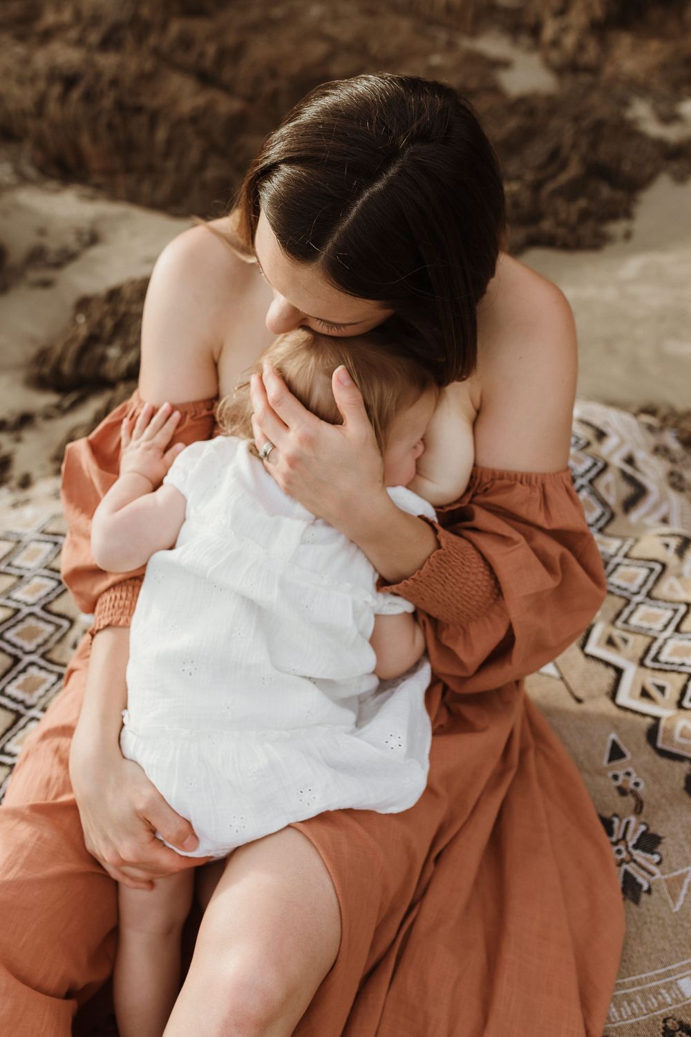 Mother in rust dress breastfeeding infant on patterned blanket outdoors at beach.