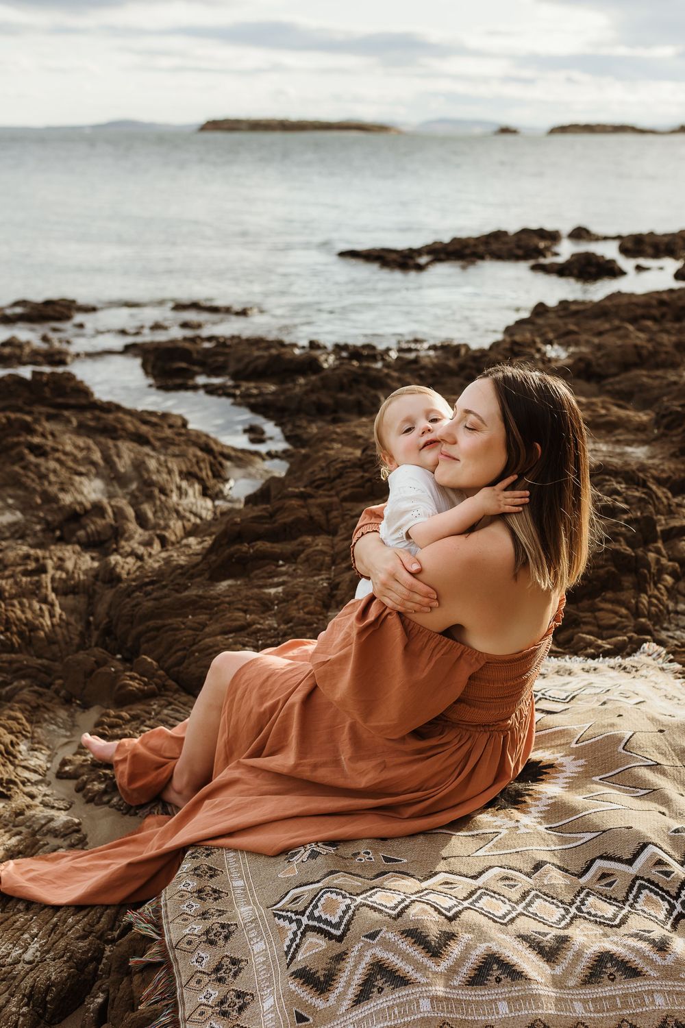 A mother in a rust dress holds her baby on a rocky beach blanket by the ocean.
