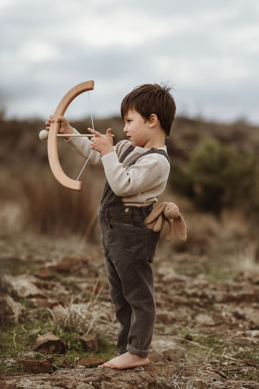 Young boy playing with wooden bow and arrow toy outdoors, wearing suspenders and barefoot in nature.