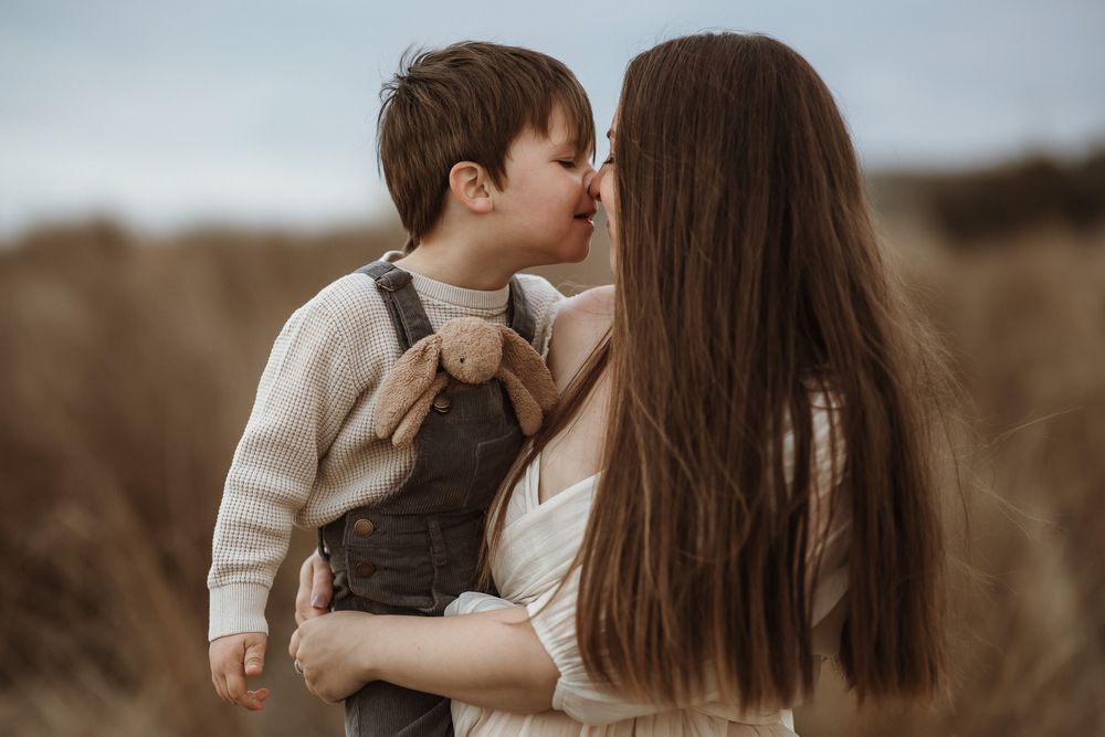 A mother holds her young son in a field, sharing a tender nose-to-nose moment outdoors.