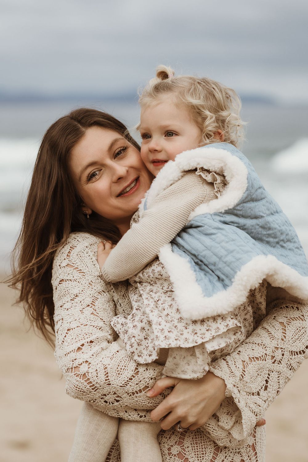A smiling mother holds her young blonde toddler on a cloudy beach, both wearing cozy outfits.