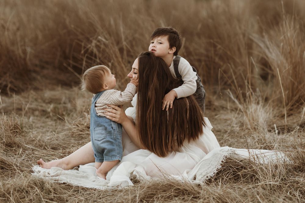 A mother sits on a white blanket in golden grass fields, embracing her two young children in a tender outdoor moment.