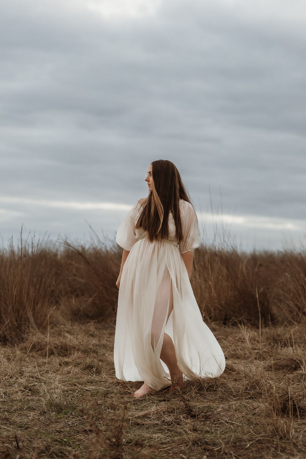 A woman in a flowing white dress stands barefoot in a dry grass field under a cloudy sky.