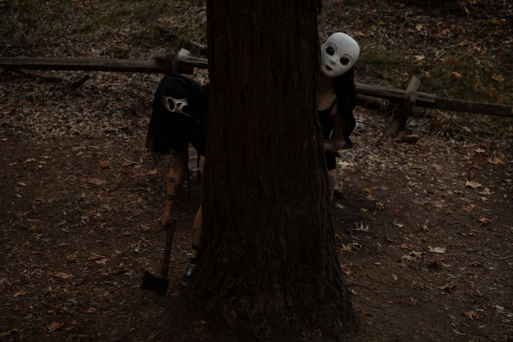 Shadowy figures lurk behind a large tree trunk in a dark forest setting.