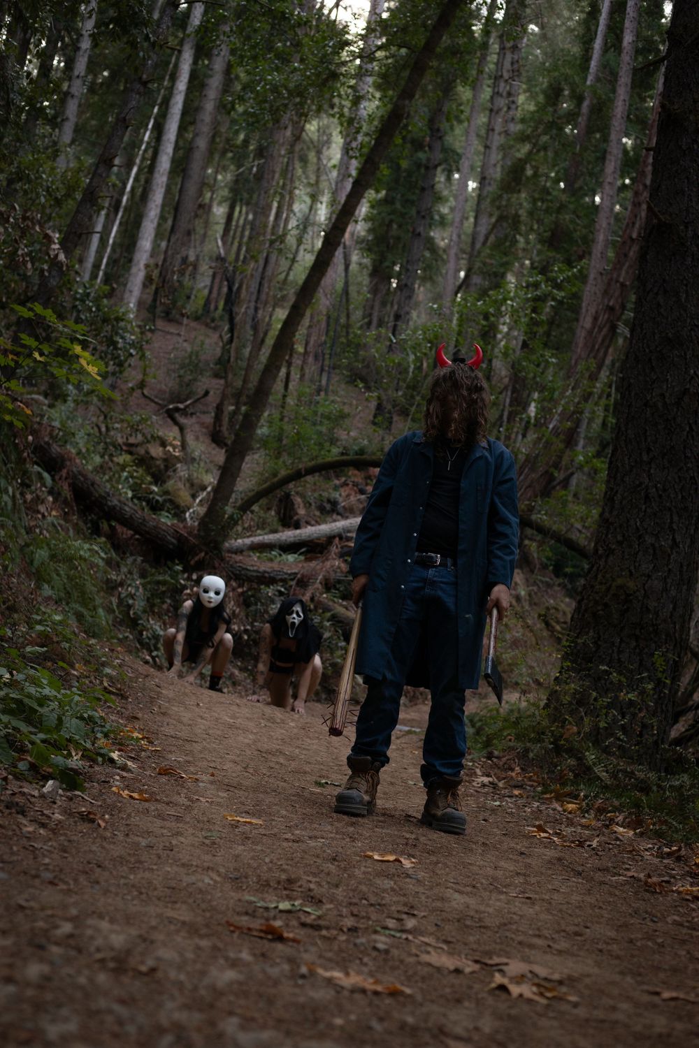 A masked figure stands on a forest trail with others approaching from behind.