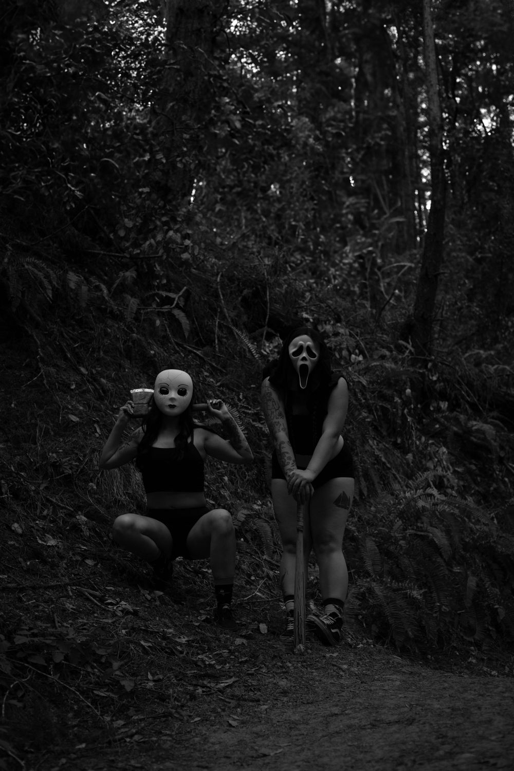 Two masked figures pose on a dirt trail in a moody forest setting.