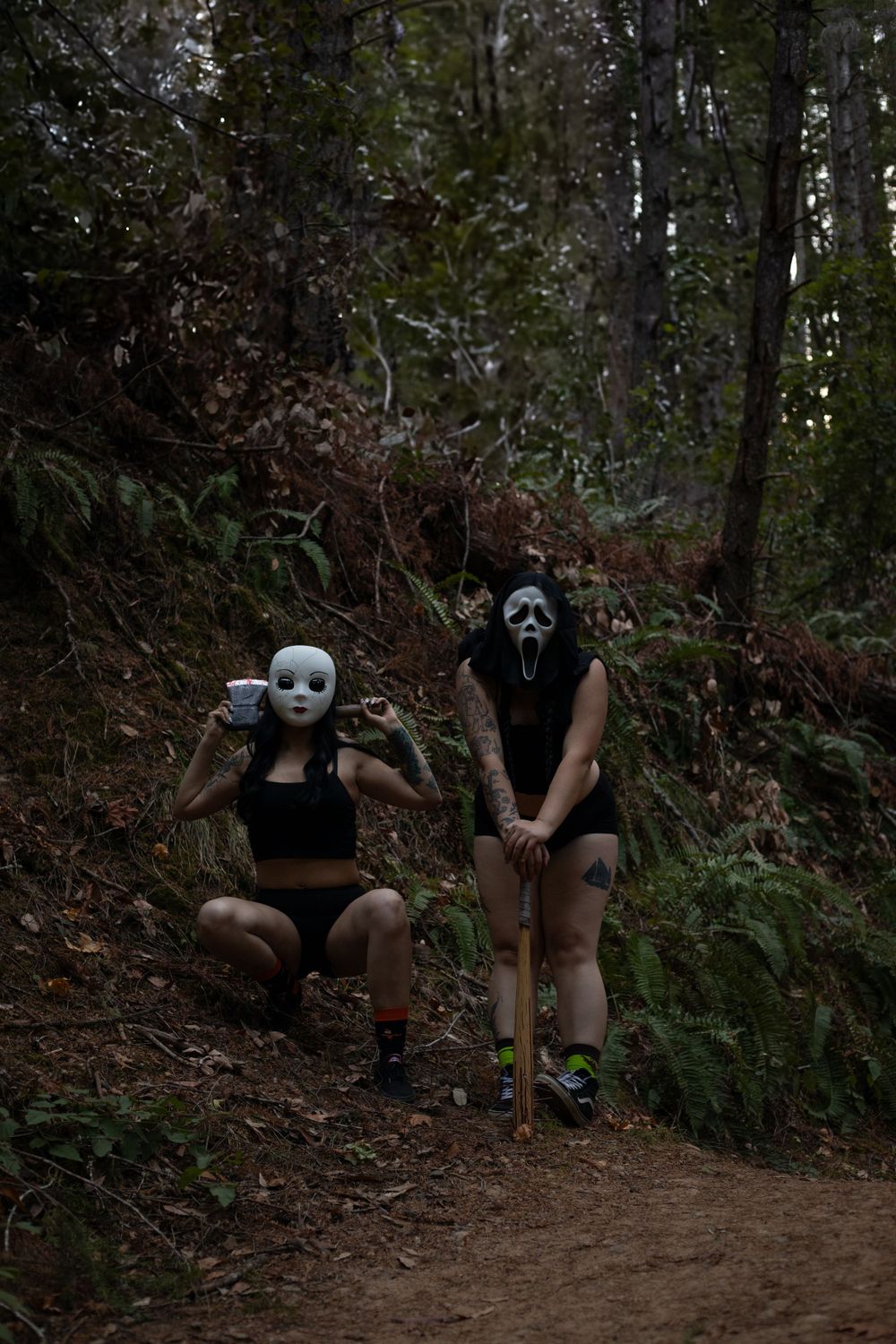 Two masked figures pose on a dirt trail in a moody forest setting.