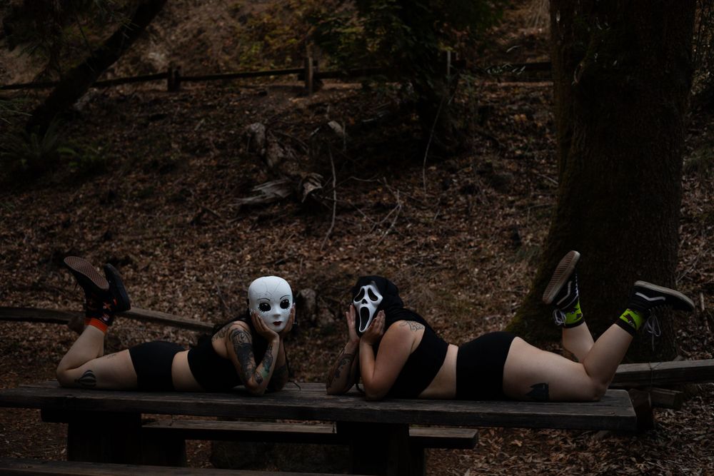 Three people wearing masks lie on a wooden bench in a forest setting.