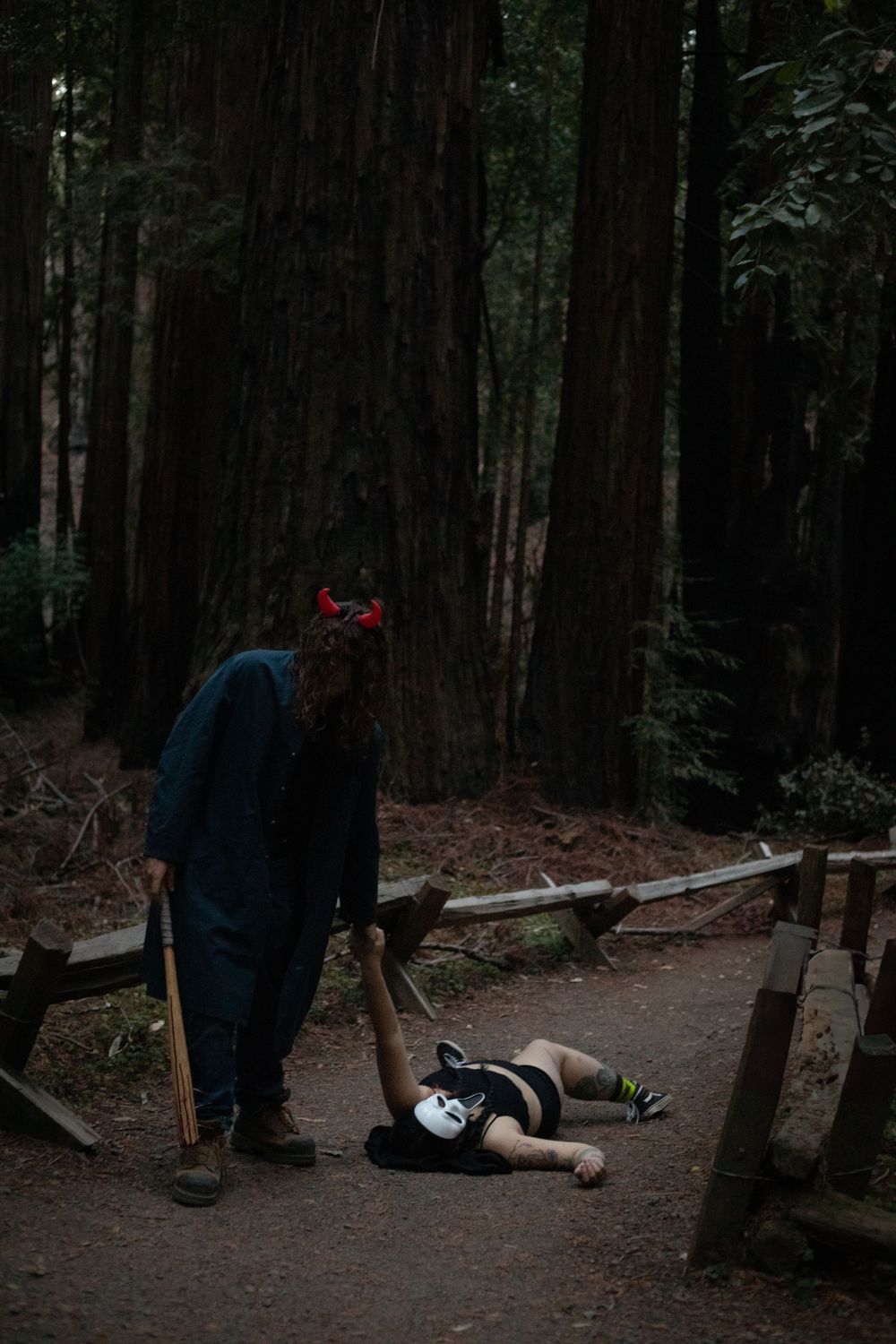 Masked figures stage an eerie scene on a forest path among tall trees.