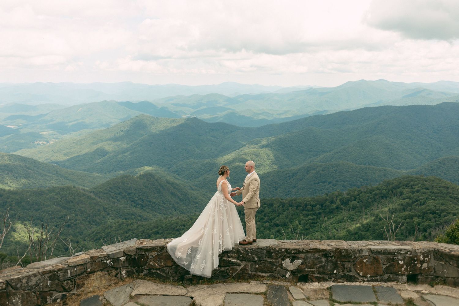 Wedding couple stands on mountain overlook with sweeping valley views below.
