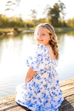 A teen in a blue and white floral dress sits on a dock by a lake during golden hour.