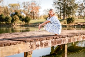 A teen in a white dress jumps playfully on a wooden dock by a serene pond during golden hour.