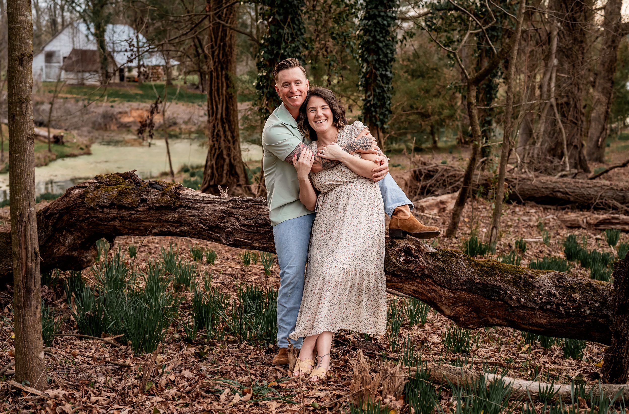 A couple embraces in a romantic forest setting with fallen logs and early spring foliage.