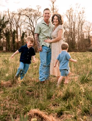 A rustic family portrait in a golden meadow at sunset shows people embracing while children play in the grass.