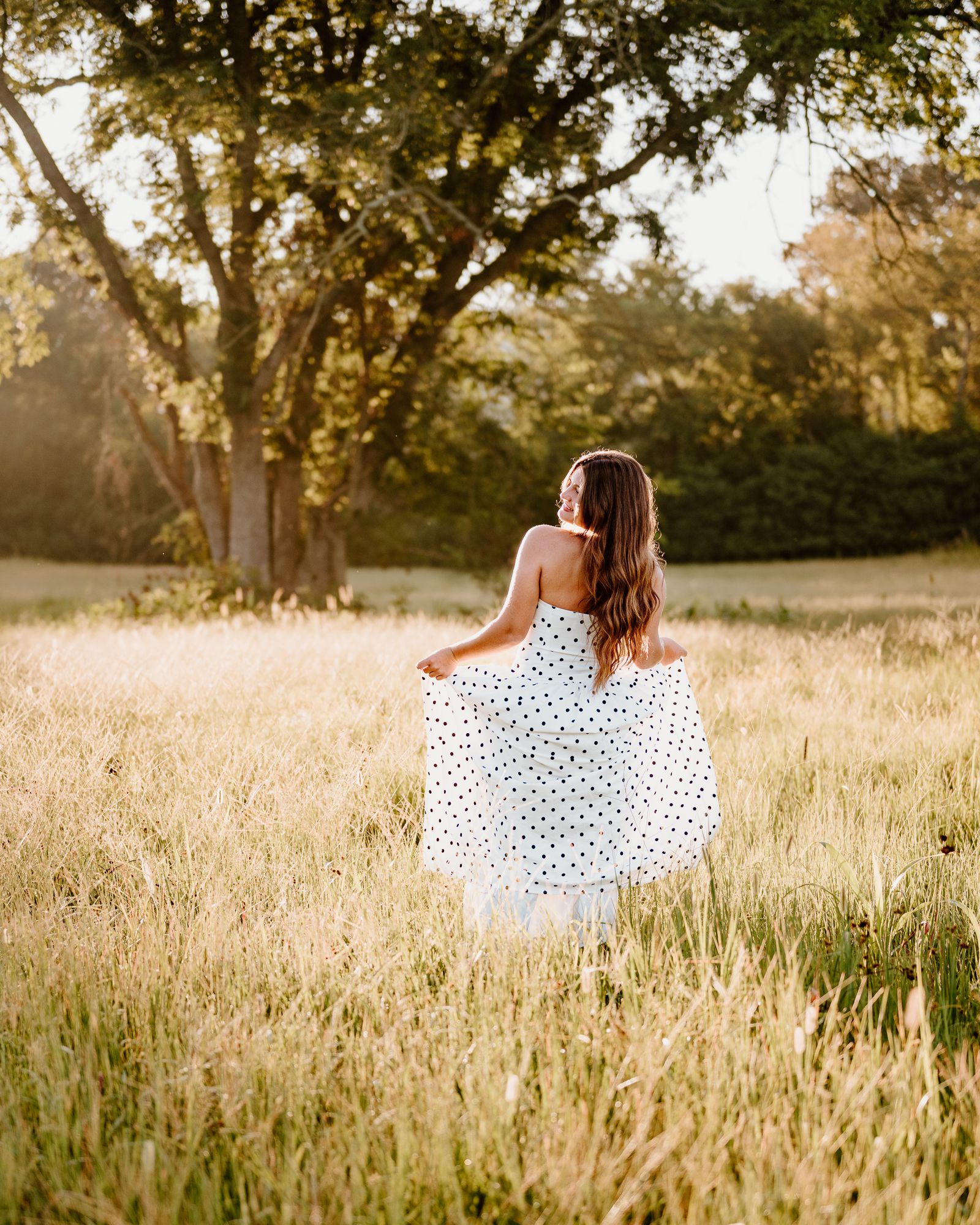 A person in a white polka dot dress walking through a golden field at sunset with trees in the background.