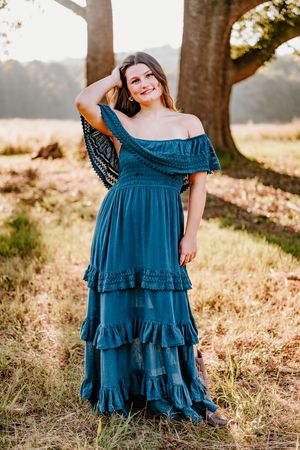 A flowy teal off-shoulder maxi dress with tiered ruffles photographed in a sunlit outdoor field during golden hour.