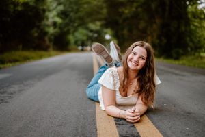 Woman in white laying on road while holding coffee and smiling at camera during outdoor photoshoot.